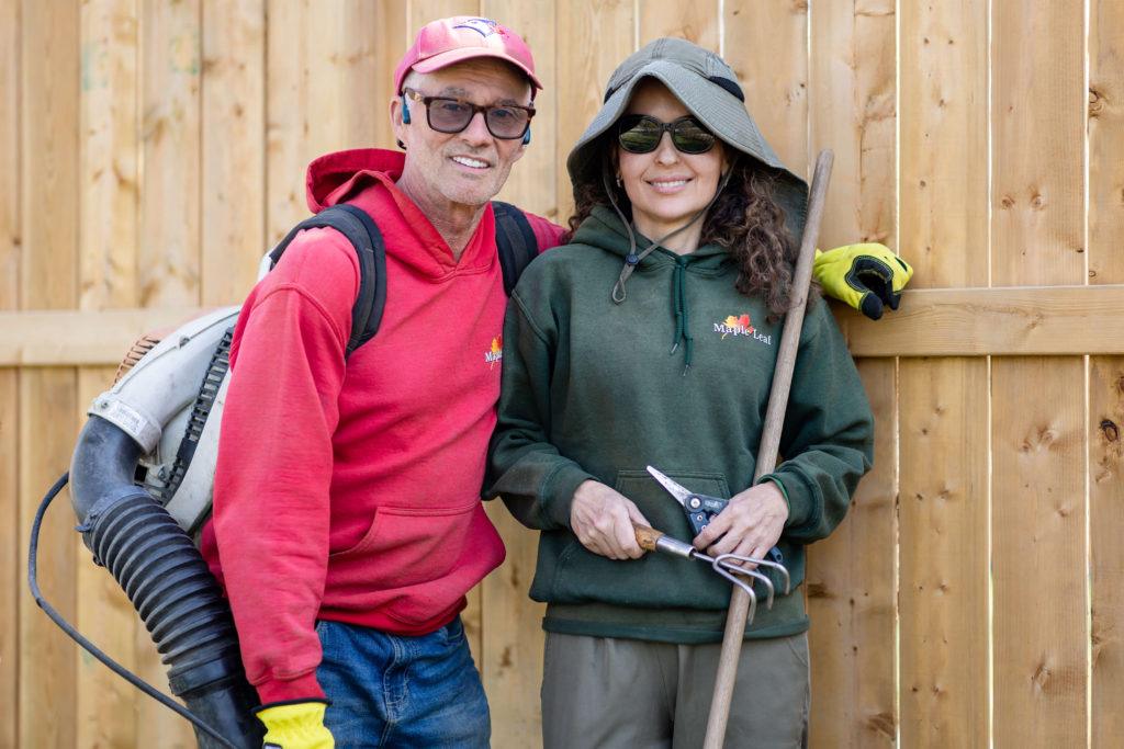 Mark and Maria, owners of Maple Leaf Lawn Maintenance in Kitchener, holding gardening tools and wearing branded clothing